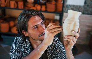 Der junge Keramiker hält seinen frischen handgefertigten Topf und macht mit einem Spezialwerkzeugstock etwas Detaillierung foto