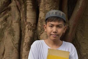Portrait asiatischer muslimischer oder islamischer Junge, der allein vor dem großen Baum steht und Buch auf der Brust hält, weicher und selektiver Fokus. foto