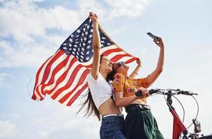 zwei patriotische fröhliche frauen mit fahrrad und usa-flagge in den händen machen selfie foto