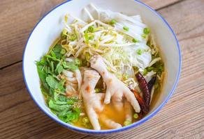 Reisnudeln mit Hühnerfüßen auf Suppenschüssel auf Holztisch Lebensmittelhintergrund - thailändisches Essen thailändische Reisnudeln würzige Currysuppe mit frischem Gemüse foto