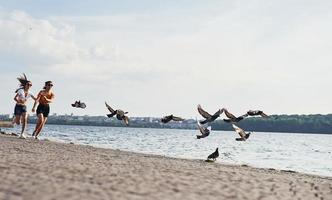 Vögel fliegen in die Luft. Zwei Freundinnen rennen und haben Spaß am Strand in der Nähe des Sees bei sonnigem Tag foto