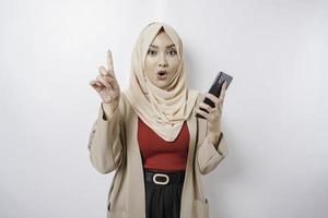 schockierte asiatische frau, die hijab trägt und auf den kopierraum auf ihr zeigt, während sie ihr telefon hält, isoliert durch weißen hintergrund foto