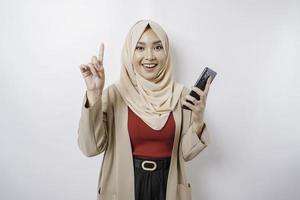 Aufgeregte asiatische Frau, die Hijab trägt und auf den Kopierbereich auf ihr zeigt, während sie ihr Telefon hält, isoliert durch weißen Hintergrund foto