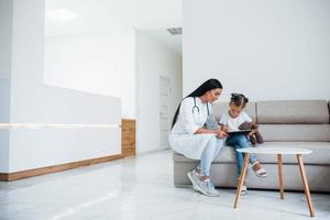 Ärztin mit Notizblock sitzt mit kleinem Mädchen in der Klinik foto