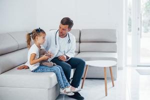 männlicher arzt in weißer uniform sitzt mit kleinem mädchen in der klinik foto