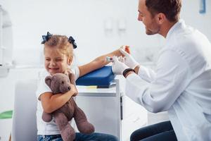 Blutprobe. Kleines Mädchen mit ihrem Teddybär ist mit Arzt in der Klinik foto