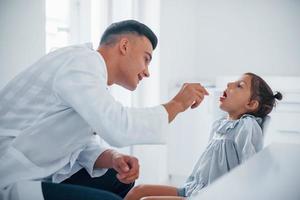 verwendet Tonsillen, um den Hals zu überprüfen. junger kinderarzt arbeitet mit kleiner weiblicher besucherin in der klinik foto