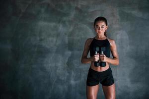 junge fitnessfrau ist im fitnessstudio in der nähe der wand mit hanteln in den händen foto