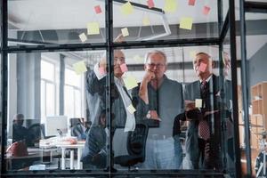 funktioniert mit Aufklebern auf dem Glas. Ein älteres Team älterer Geschäftsmannarchitekten trifft sich im Büro foto