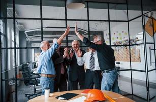 Arbeit gut gemacht, gibt sich gegenseitig High Five. Ein älteres Team älterer Geschäftsmannarchitekten steht zusammen im Büro foto