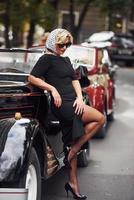 blonde Frau mit Sonnenbrille und in schwarzem Kleid, die sich an alte Oldtimer anlehnt foto