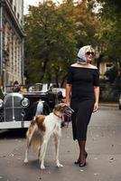 Blonde Frau mit Sonnenbrille und schwarzem Kleid in der Nähe eines alten Oldtimers mit ihrem Hund foto