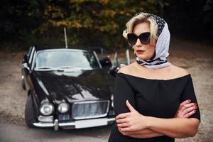 Blonde Frau mit Sonnenbrille und schwarzem Kleid in der Nähe eines alten Oldtimers foto