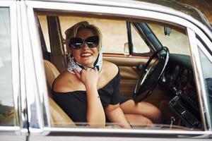 Blonde Frau mit Sonnenbrille und schwarzem Kleid sitzt in einem alten Oldtimer foto