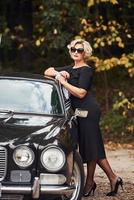 Blonde Frau mit Sonnenbrille und schwarzem Kleid in der Nähe eines alten Oldtimers foto