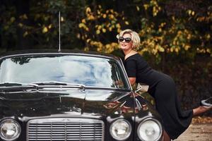 Blonde Frau mit Sonnenbrille und schwarzem Kleid in der Nähe eines alten Oldtimers foto