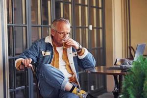 Stylischer Senior in modischer Kleidung und Brille sitzt mit einem Getränk im Café foto