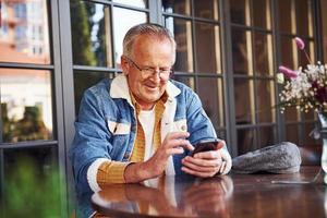 Stylischer Senior in modischer Kleidung und Brille sitzt im Café mit Telefon foto