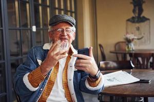 Stylischer Senior in modischer Kleidung und Brille sitzt mit Zigarette und Telefon im Café foto