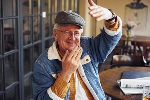 Stylischer Senior in modischer Kleidung und Brille sitzt mit Zigarette und Gesprächen im Café foto
