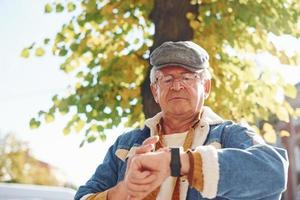 Stylischer Senior in modischer Kleidung und mit Brille steht in der Stadt und kontrolliert die Zeit auf seiner Uhr foto