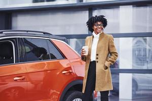 junge afroamerikanerin in gläsern und mit einer tasse getränk steht draußen in der nähe eines modernen autos foto