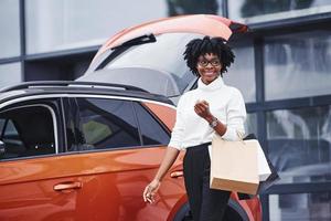 junge afroamerikanerin mit brille steht nach dem einkauf mit paketen im freien in der nähe eines modernen autos foto