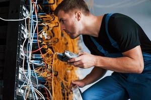 Junger Mann in Uniform mit Messgerät arbeitet mit Internetgeräten und Kabeln im Serverraum foto