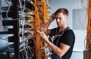 Der junge Mann in Uniform fühlt sich verwirrt und sucht nach einer Lösung mit Internetgeräten und Kabeln im Serverraum foto