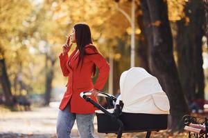 mutter im roten mantel geht zur herbstzeit mit ihrem kind im kinderwagen im park spazieren und raucht foto