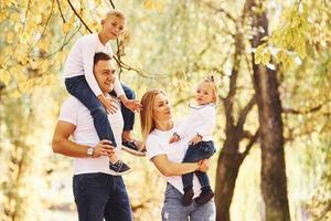 mutter und vater halten kinder an den schultern und in den händen. Fröhliche junge Familie macht zusammen einen Spaziergang in einem Herbstpark foto