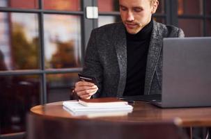 Eleganter junger Mann in formeller Kleidung sitzt mit seinem Laptop und seiner Kreditkarte in der Hand im Café foto