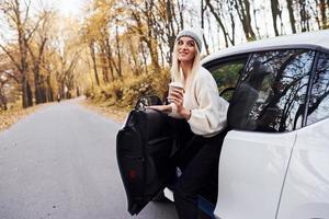 mit Tasse Getränk in den Händen. Mädchen haben Herbstausflug mit dem Auto. modernes, brandneues Auto im Wald foto