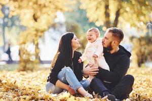 auf dem Boden sitzen. Fröhliche Familie, die sich zusammen mit ihrem Kind im schönen Herbstpark amüsiert foto