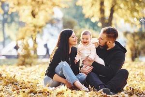auf dem Boden sitzen. Fröhliche Familie, die sich zusammen mit ihrem Kind im schönen Herbstpark amüsiert