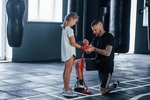 beim Tragen der Bandagen helfen. Der junge tätowierte Boxtrainer unterrichtet ein süßes kleines Mädchen im Fitnessstudio
