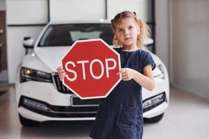 Porträt eines süßen kleinen Mädchens, das im Autosalon ein Straßenschild in den Händen hält foto