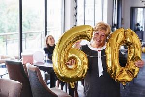 mit Ballons der Nummer 60 in den Händen. ältere frau mit familie und freunden, die drinnen einen geburtstag feiern foto