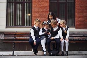 Schulkinder in Uniform, die mit Smartphone draußen auf der Bank sitzen foto