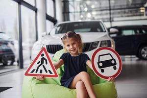 Porträt eines süßen kleinen Mädchens, das Verkehrsschilder im Automobilsalon in den Händen hält foto