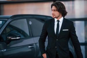 Porträt eines hübschen jungen Geschäftsmannes in schwarzem Anzug und Krawatte im Freien in der Nähe eines modernen Autos foto