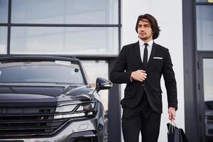 Porträt eines hübschen jungen Geschäftsmannes in schwarzem Anzug und Krawatte im Freien in der Nähe eines modernen Autos und mit Einkaufstüten foto