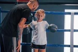 auf dem Boxring stehen. Der junge tätowierte Boxtrainer unterrichtet ein süßes kleines Mädchen im Fitnessstudio foto