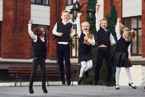 gruppe von kindern in schuluniform springen und spaß im freien zusammen in der nähe des bildungsgebäudes foto
