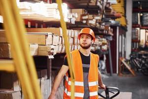 junger männlicher arbeiter in uniform ist im lager und schiebt hubwagen foto