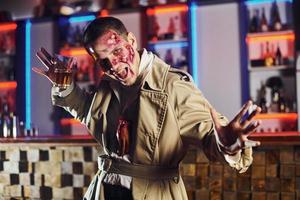 mit Getränk in der Hand. Porträt des Mannes, der auf der thematischen Halloween-Party in Zombie-Make-up und Kostüm ist foto
