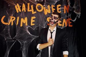 Porträt eines Mannes, der auf der thematischen Halloween-Party in gruseligem Skelett-Make-up und Kostüm gegen die Wand des Tatorts ist foto