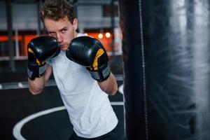junger mann in weißem hemd und boxschutzhandschuhen, die übungen im fitnessstudio mit schubsack machen foto