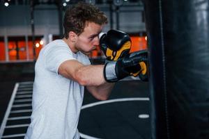 junger mann in weißem hemd und boxschutzhandschuhen, die übungen im fitnessstudio mit schubsack machen foto