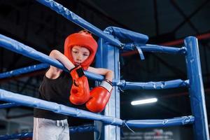 Müder Junge in Schutzausrüstung, der sich auf die Knoten des Boxrings stützt foto